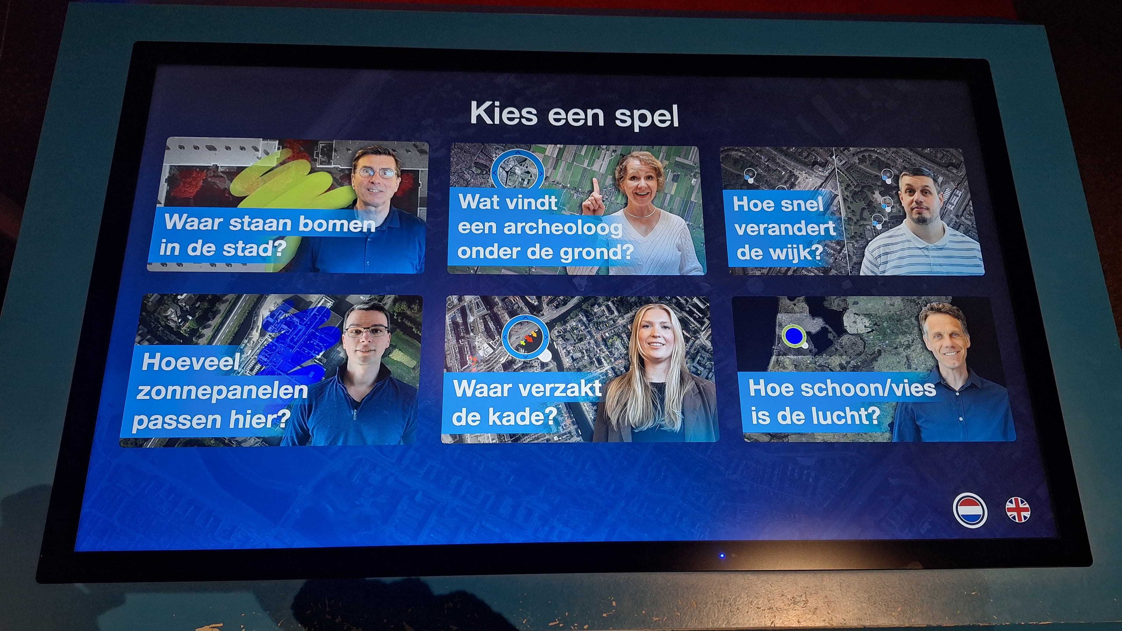 Foto van het interactieve scherm in de tentoonstelling met daarop afbeeldingen van de zes experts die satellietdata gebruiken in hun werk.